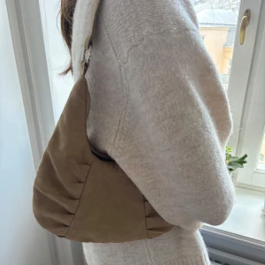 Beige axelväska i mocka - Snygg beige axelväska i mjukt mockamaterial. Väskan har en stilren design med rynkade detaljer och en axelrem med dekorativa ringar. Perfekt för att ge din outfit en trendig touch.
