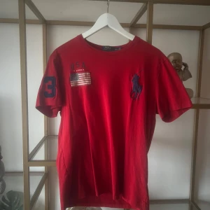 Röd t-shirt från Ralph Lauren - Säljer en röd t-shirt från Ralph Lauren med USA-flagga och nummer 3 på ärmen. Den har en klassisk passform och är kortärmad. Perfekt för en sportig look.