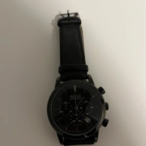 Svart klocka från Hugo Boss - Snygg svart klocka från Hugo Boss med läderarmband. Klockan har en stilren urtavla med flera funktioner och en robust design. Perfekt för den som gillar en klassisk och tidlös stil.