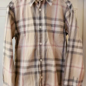 Burberry skjorta - Burberryskjorta i klassiskt rutmönster i milda färger (beige, svart, vitt, rött). Kan bäras långärmad, men man kan även vika upp ärmarna och fästa med en flärp och knapp, se bild.  Väldigt bra skick, ser nästan ny ut utan synliga defekter. Extraknappen sitter kvar.  Storlek 12 år, 152 cm. Passar både killar och tjejer.