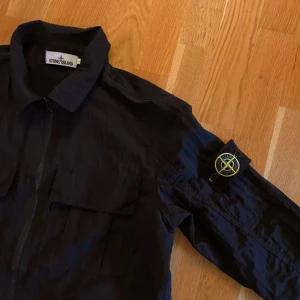 Svart overshirt från Stone Island - Säljer en stilren svart overshirt från Stone Island med dragkedja och ikonisk logga på ärmen. Perfekt för lager-på-lager och har en klassisk krage. Passar bra till både casual och mer uppklädda tillfällen.perfekt till våren då det är ganska tunt material                  ⚠️ej äkta⚠️