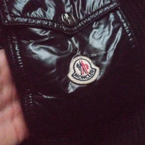 Svart dunväst från Moncler - Snygg svart dunväst från Moncler med glansigt material och huva. Västen har knappar och dragkedja framtill samt en broderad logotyp på bröstet. Perfekt för kyliga dagar när du vill hålla stilen.