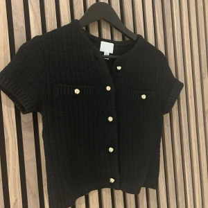 Svart stickad kofta med guldknappar - Snygg svart stickad kofta med korta ärmar och eleganta guldknappar framtill. Den har en ribbad struktur och två dekorativa fickor på bröstet. Perfekt för en stilren look.