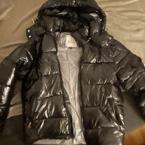 Svart pufferjacka från Moncler - Säljer en snygg svart pufferjacka från Moncler med glansig finish. Jackan har en huva och dragkedja framtill. Perfekt för kyliga dagar och ger en stilren look. Moncler-logotypen är broderad på bröstet.