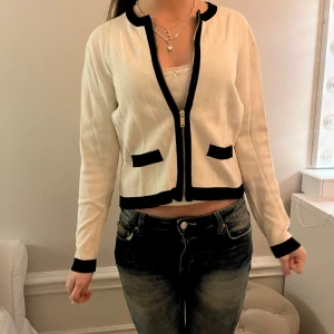 Vit och svart kofta med dragkedja - Snygg vit kofta med svarta detaljer och dragkedja framtill. Den har långa ärmar och två dekorativa fickor. Perfekt för en stilren look.