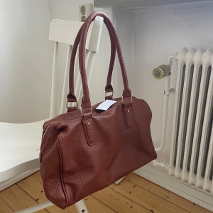 Brun axelväska i skinn - Snygg brun axelväska i skinn med två  handtag, dragkedja upptill och flera fack. Perfekt till vardags eller till skola/jobb då den tex rymmer dator. Köpt second hand från Erikshjälpen därav okänt märke.