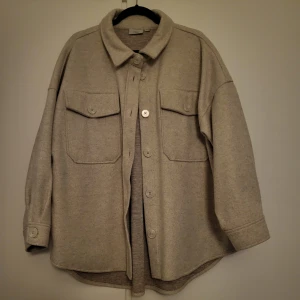 Beige overshirt med knappar - Säljer en stilren beige overshirt med knappar framtill. Den har två stora bröstfickor med lock och knappar, samt en klassisk krage. Perfekt för lager-på-lager-stil och passar till många olika outfits.