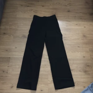 Svarta byxor från Madlady - Snygga svarta byxor från Madlady med hög midja och en rak passform. Perfekta för en stilren look. Byxorna har en enkel design utan mönster eller extra detaljer.