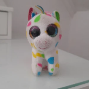 Flerfärgad enhörningsnyckelring från Ty - Söt enhörningsnyckelring från Ty med stora glittriga ögon. Den är vit med färgglada prickar i rosa, blått, grönt och gult. Har en rosa man och svans samt en praktisk metallklämma för att fästa på väskan eller nyckelknippan.