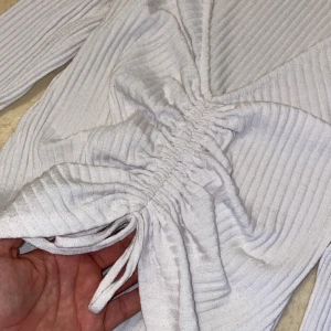 Vit ribbad topp med knytning - Vit ribbad topp med lång ärm och knytning framtill. Toppen har en v-ringad design och är reglerbar i knytningen. 