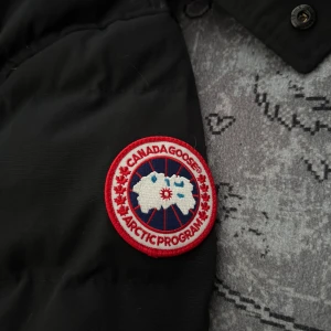 Svart dunjacka från Canada Goose - Säljer en svart dunjacka från Canada Goose med ikonisk logga på ärmen. Jackan har en hög krage och knäppning med både dragkedja och knappar. Perfekt för kalla vinterdagar med sin varma och skyddande design. 🖤via intresse skriv till mig så skickar jag mer bilder 