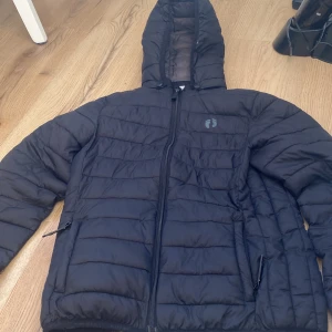 Svart pufferjacka med huva - Säljer en svart pufferjacka med huva och dragkedja framtill. Jackan har långa ärmar och är perfekt för kyligare väder. Den har en stilren design med quiltade detaljer och praktiska fickor med dragkedja.