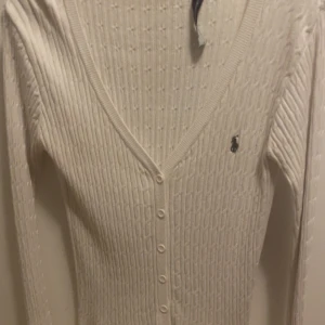 Vit ribbad kofta från Polo Ralph Lauren - Säljer en stilren vit ribbad kofta från Polo Ralph Lauren. Koftan har en v-ringad design med knappar framtill och det klassiska logotypbroderiet på bröstet. Perfekt för en elegant och avslappnad look. Ända defekten är att etiketten har lossnat av på ena sidan