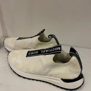 Vita sneakers från Michael Kors - Snygga vita sneakers från Michael Kors med svart logotypband över ovansidan. Skorna har en stilren design med en bekväm sula och diskret mönster. Perfekta för en trendig look.