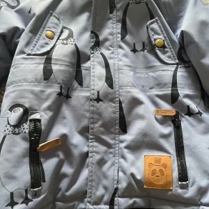 Grå barnjacka med pingvinmönster från Mini Rodini - Säljer en superfin grå barnjacka från Mini Rodini med gulligt pingvinmönster. Jackan har en mysig huva med fuskpäls och praktiska fickor med dragkedjor och knappar. Perfekt för kyliga dagar! Jackan är endast testad men notera att det står initialer inuti jackan detta var då vi trodde jackan skulle användas på förskola🥰