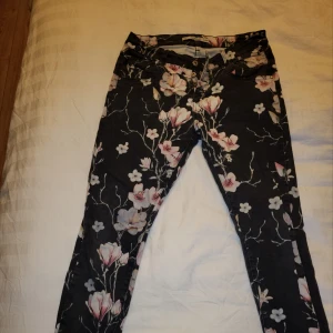 Blommiga byxor med svart bas - Snygga byxor med ett blommigt mönster på en svart bas. Byxorna har en normal passform och är perfekta för att ge en färgglad touch till din outfit. De har knappar framtill.