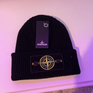 Svart ribbad mössa från Stone Island - Frakar samma dag!                                          Snygg svart ribbad mössa från Stone Island med det ikoniska broderade märket framtill. Perfekt för att hålla värmen med stil. Mössan är i ett klassiskt och tidlöst utförande.