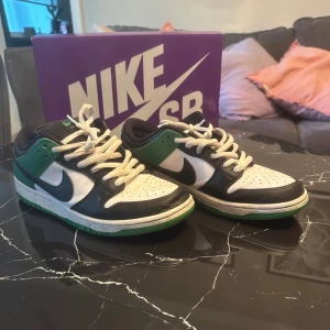 Nike SB Dunk i svart, vit och grön - Snygga Nike SB Dunk sneakers i en cool färgkombination av svart, vit och grön. Skorna har klassisk snörning och en robust sula för extra grepp. Perfekta för både skate och vardag.