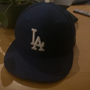 Blå keps från New Era - Snygg blå keps från New Era med LA-logga framtill. Kepsen har en klassisk design med böjd skärm och är tillverkad i polyester. Perfekt för att ge en sportig touch till din outfit.