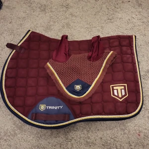 Vinrött schabrak och huva från Trinity - Säljer ett snyggt vinrött schabrak med matchande huva från Trinity. Schabraket har en quiltad design med blå och guldiga detaljer samt Trinity-loggan. Huvan är i samma vinröda färg med gudigl kant. Passar så fint oavsett vilken färg hästen har på pälsen. Dock är det lite slitet