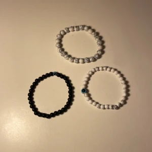 Tre elastiska armband med pärlor - Säljer tre elastiska armband med pärlor. Två av armbanden är vita med ett blått öga som detalj, och ett är svart med samma blå öga. Perfekta för att bära tillsammans eller var för sig.