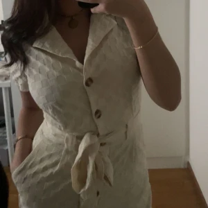 Beige playsuit med knappar - Säljer en stilren beige playsuit med knappar framtill och ett knytband i midjan. Playsuiten har korta ärmar och ett diskret mönster som ger en elegant touch. Perfekt för en somrig look! använd 1 gång!