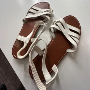 Vita sandaler med remmar - Snygga vita sandaler med flera remmar och en bekväm brun sula. Perfekta för varma sommardagar och ger en stilren look. Remmarna ger bra stöd och komfort.