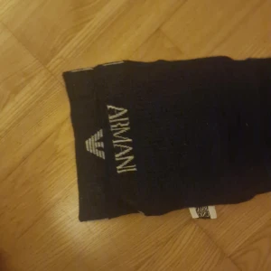 Svart halsduk från Armani - Snygg svart halsduk från Armani med logotyp i grått. Perfekt för att hålla sig varm och stilren under kyliga dagar. Den är lång och kan enkelt stylas på olika sätt.