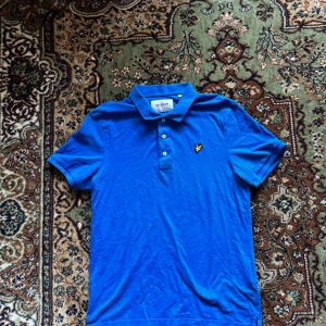 Blå pikétröja från Lyle & Scott - Säljer en snygg blå pikétröja från Lyle & Scott med klassisk krage och tre knappar. Tröjan har en gul logotyp på bröstet och är tillverkad i ekologiskt material. Perfekt för en stilren look!