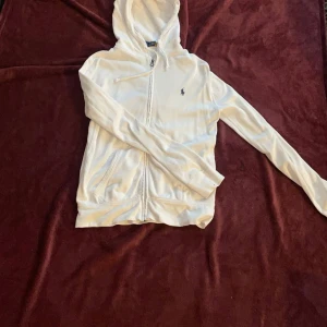 Vit hoodie från Ralph Lauren - Säljer en stilren vit hoodie från Ralph Lauren med dragkedja och den klassiska loggan broderad på bröstet. Tröjan har en justerbar huva och långa ärmar, perfekt för en avslappnad look. Inte använd mycket då den inte passade, köpt för 2300 kr men mitt pris 450. 