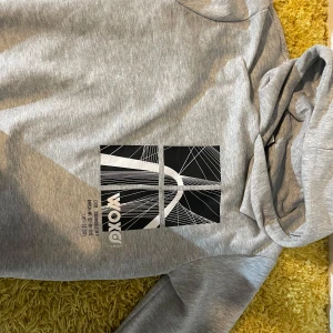 Grå hoodie från Woxon - Säljer en stilren grå hoodie från Woxon med ett coolt grafiskt tryck på framsidan. Tröjan har långa ärmar och en bekväm huva. Perfekt för en avslappnad stil.