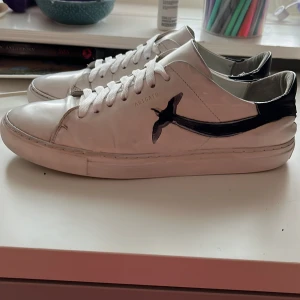 Vita skor från arigato - Snygga vita sneakers från arigato med svart detalj på sidan och hälen. Skorna har snörning och en klassisk design som passar till många stilar. Perfekta för en avslappnad look.