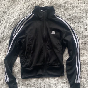 Svart träningsjacka från Adidas - Säljer en svart träningsjacka från Adidas med de klassiska tre vita ränderna längs ärmarna. Jackan har dragkedja framtill och en hög krage. Bra skick🌟