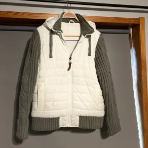 Cardigan jacka med stickade armar - Snygg vit och grå jacka med quiltad design och stickade ärmar. Jackan har en dragkedja framtill och en mysig huva med dragsko. Perfekt för kyliga dagar när du vill hålla stilen. 🧥