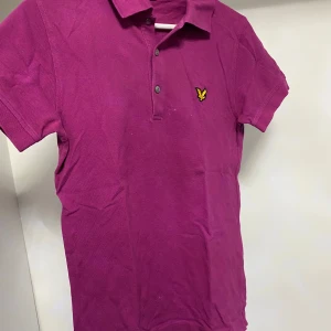 Lila pikétröja från Lyle & Scott - Säljer en snygg lila pikétröja från Lyle & Scott med klassisk krage och knappslå. Tröjan har korta ärmar och det ikoniska logotypmärket på bröstet. Perfekt för en stilren look!
