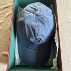 Blå keps från Loro Piana - Snygg blå keps från Loro Piana med broderad logotyp framtill. Kepsen har en klassisk design med rundad skärm och justerbar passform. Insidan har ett stilrent rutigt mönster.