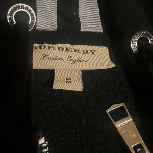 Svart hoodie från Burberry - Säljer en stilren svart hoodie från Burberry med dragkedja och klassiskt rutigt mönster på insidan av huvan. Jackan har långa ärmar och en broderad logotyp på bröstet. Perfekt för en casual look.
