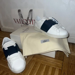 Valentino Garavani  - Snygga Valentino Garavani sneakers i vitt med en bred blå rem över ovansidan. Skorna har en klassisk design med snörning och är tillverkade i skinn. Perfekta för en stilren look. Fått nyligen fast passade tvyär inte. Nypris 7.899kr