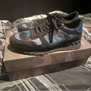 Camouflage sneakers från Valentino - Snygga sneakers från Valentino i ett coolt blått och svart camouflage-mönster. Skorna har snörning och är perfekta för en trendig look. De kommer i originalförpackning med extra skosnören.