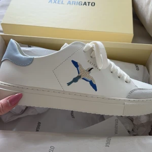 Vita sneakers från Axel Arigato - Snygga vita sneakers från Axel Arigato med en broderad fågel i blått. Skorna har ljusblå detaljer vid hälen och klassisk snörning framtill. Köpa på deras hemsida April 2024. Mycket bra skick. Nypris: 2550. Säljer för 1000kr storlek 38. Box kommer med skorna! 💙💙💙