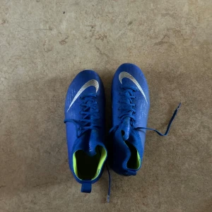 Blå fotbollsskor från Nike - Snygga blå fotbollsskor från Nike med vit swoosh-logga. Skorna har snörning och en hög krage för extra stöd. Perfekta för fotbollsplanen med sina dobbar för bra grepp.