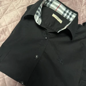 Burberry skjorta - Snygg svart skjorta från Burberry med klassiskt rutigt mönster på insidan av kragen. Skjortan har långa ärmar och knappar framtill. Perfekt för en stilren look. Skjortan är i storlek M Men är väldigt liten i storlek så sitter som S!!!