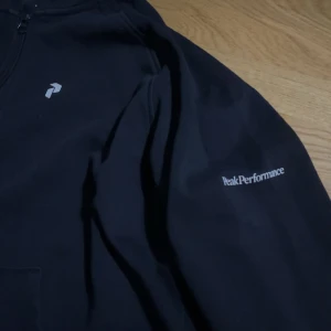 Svart hoodie från Peak Performance - Säljer en svart hoodie från Peak Performance i storlek XL. Den har en dragkedja framtill och en klassisk huva. Perfekt för både vardag och träning. Märket är diskret tryckt på bröstet och ärmen. Superbekväm och stilren!