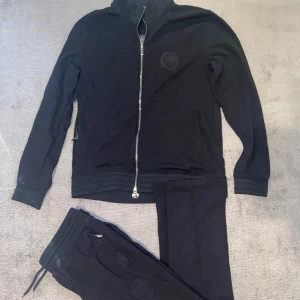 Svart tröja med dragkedja från Stefano Ricci - Säljer en stilren svart tröja från Stefano Ricci i storlek XXL men som är liten i strlk och passar även L. Tröjan har en dragkedja framtill och ett diskret broderat emblem på bröstet. Perfekt för en avslappnad men ändå elegant look. 