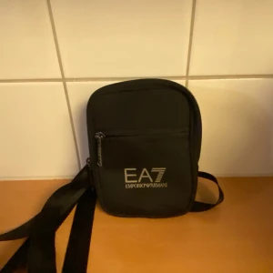 Svart axelväska från Emporio Armani - Säljer en stilren svart axelväska från Emporio Armani. Väskan har en justerbar axelrem och en praktisk dragkedja framtill. Perfekt för att bära med sig det viktigaste på ett snyggt sätt. Passar både till vardags och fest!