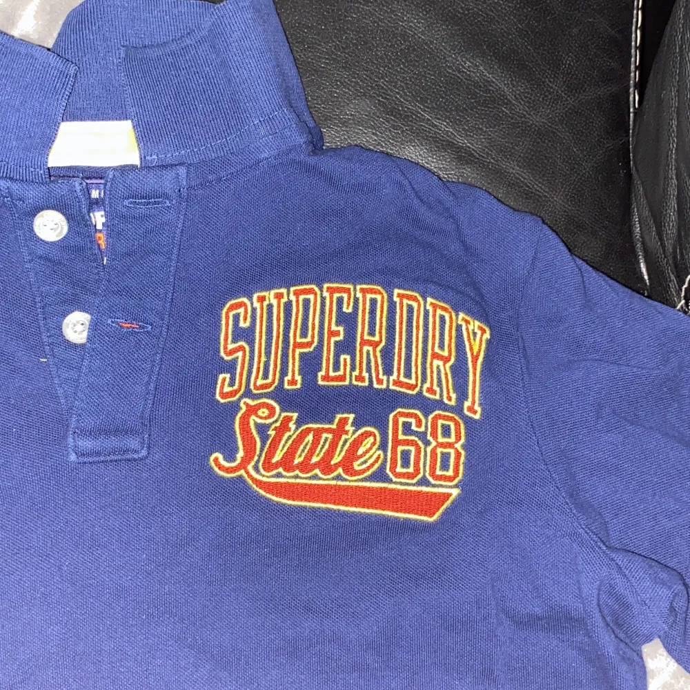 Superdry Premium Polo i storlek medium, mörkblå färg. I mycket gott skick, sparsamt använd och utan synliga skador eller fläckar. Klassisk stil med broderade detaljer och tryck på framsidan samt ärmen. . T-shirts.