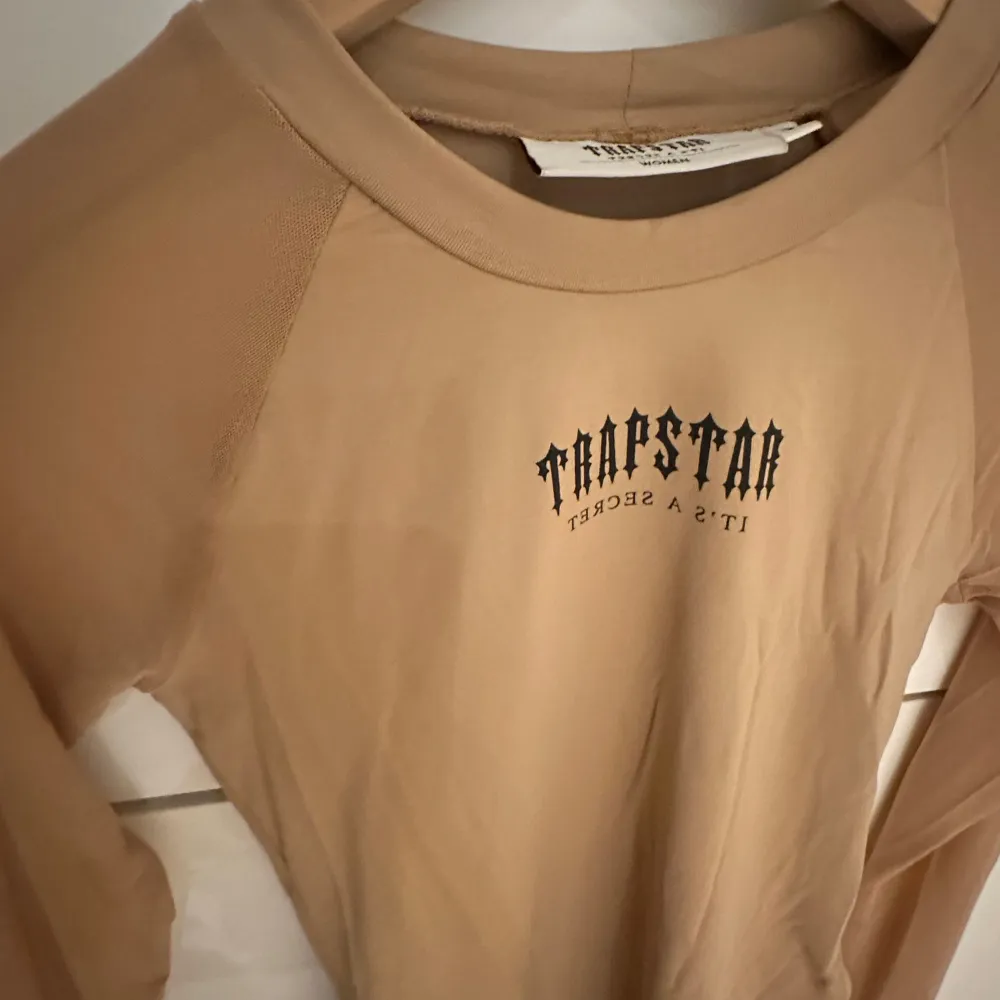 Säljer en beige långärmad body från Trapstar med trycket 'It's a Secret' på bröstet. Den är tight och formar kroppen. Materialet är mjukt och stretchigt. Använd 2 gånger. Den har så att man kan lägga in tummarna så syns liggan på händerna.. Toppar.