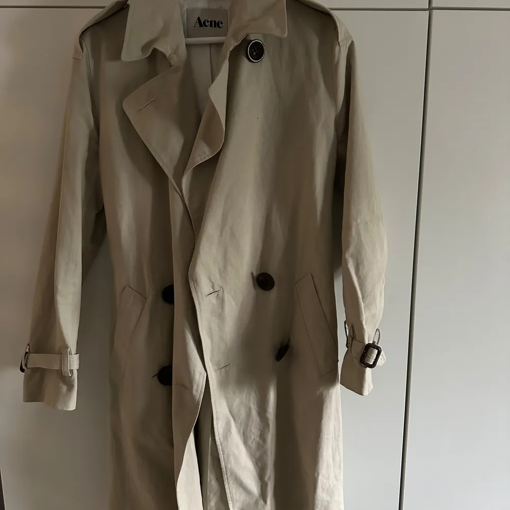 Säljer en snygg beige trenchcoat från Acne i storlek 34. Den är gjord av 73% bomull och 27% linne, vilket gör den perfekt för vår och höst. Jackan har långa ärmar och knappar framtill, samt ett bälte i midjan för en stilren look. Perfekt för att hålla stilen när det är lite kyligare ute!. Jackor.