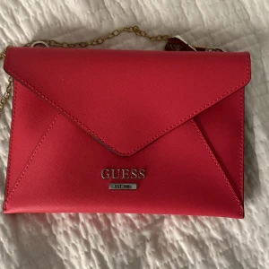 Röd kuvertväska från Guess - Säljer en snygg röd kuvertväska från Guess. Väskan har en elegant design med en guldfärgad kedja och Guess-logga framtill. Perfekt för en utekväll eller fest! Den har en praktisk dragkedja för säker förvaring av dina saker. 