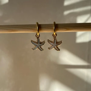 Guldörhängen med sjöstjärna - Säljer ett par superfina guldörhängen med hängande sjöstjärnor. De är perfekta för att ge en somrig touch till din outfit. Sjöstjärnorna har en fin detaljering som gör dem unika och de hänger från en enkel guldring. Helt nya! Handgjorda 